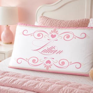 May include: A rectangular white pillowcase featuring a pink decorative border with scrollwork and heart motifs. The name Lillian is printed in a cursive pink script in the centre of the pillowcase, framed by symmetrical pink flourishes and floral accents.