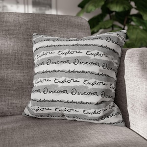 Puede incluir: Cojín cuadrado con un diseño de rayas grises y blancas. Las palabras "Explore", "Discover" y "Adventure" están escritas en una fuente manuscrita. El cojín está sobre un sofá gris.