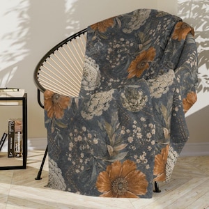 May include: A soft throw blanket featuring a dark slate blue background with a repeating botanical pattern of large rust-colored flowers, delicate white lace-like floral clusters, and small green leaves draped over a black wire frame chair.