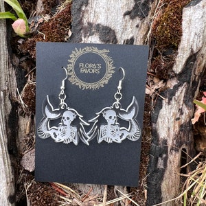 May include: A pair of silver skeleton mermaid earrings on a black card with the text "Flora's Favors".