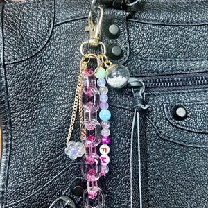 May include: A gold-toned keychain with a lobster clasp, attached to a black leather bag. The keychain features a pink chain, a gold chain, a flower charm, and a strand of colorful beads. The letters "F M" are visible on the beads.