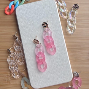 May include: Set of five pairs of colorful chunky chain earrings. The earrings are made of plastic and have a gold-tone metal finding. The earrings are displayed on a white surface.