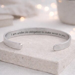 May include: A silver-colored cuff bracelet with the engraved text: "I am under no obligation to make sense to you." The open-ended bracelet is displayed on a light-colored, textured surface.