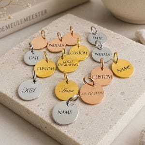Könnte beinhalten: Eine Sammlung personalisierter Metallanhänger in Gold, Silber und Roségold. Jeder Anhänger hat ein kreisförmiges Design mit eingraviertem Text, darunter Namen, Initialen, Daten und die Wörter "Custom" und "Logo Engraving".
