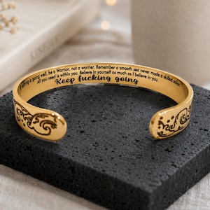 May include: Gold-colored cuff bracelet with engraved text. The bracelet features the phrase "Keep fucking going" and other motivational quotes. Decorative wave patterns are etched on the ends. The bracelet is displayed on a dark, textured surface.
