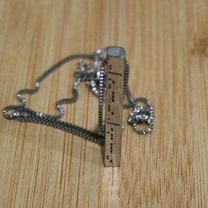 以下が含まれることがあります： 銀色のチェーンネックレスに木製のペンダントがついたもの。ペンダントには点字の点とダッシュの模様が刻まれています。