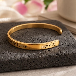 May include: A gold-coloured cuff bracelet with the engraved text "of the Future" and "prov 31:25". The bracelet is resting on a dark grey, textured surface. The bracelet is a simple, elegant design.