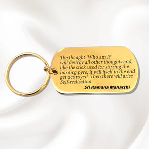 Może przedstawiać: Złoty metalowy brelok do kluczy z prostokątną zawieszką. Zawieszka zawiera wygrawerowany cytat Sri Ramany Maharshiego. Cytat jest w czarnym tekście i dotyczy samorealizacji. Brelok ma okrągły pierścień.