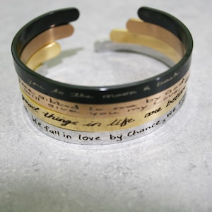 Peut inclure: Un ensemble de trois bracelets empilés avec des messages gravés. Le bracelet du haut est noir, le bracelet du milieu est doré et le bracelet du bas est argenté. Les messages sur les bracelets sont : "You to the moon & back", "Good things in life", et "We fall in love by chance we s".