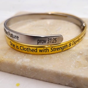 Könnte beinhalten: Ein gold- und silberfarbenes Armkettchen mit der Inschrift "She is Clothed with Strength & Dignity and she laughs at the Future" und "prov 31:25" auf der Innenseite.