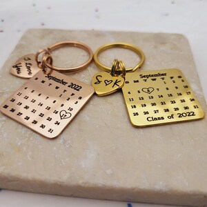 May include: Two gold keychains with engraved heart-shaped charms. One keychain features a calendar with the month of September 2022 and the number 17 circled. The other keychain has the initials "S♡K" and the text "Class of 2022".