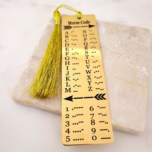 May include: A gold bookmark with a tassel features a Morse code chart for letters and numbers. The bookmark has a black arrow pointing to the right and a double arrow pointing to the left.