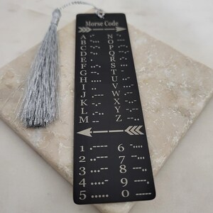 May include: A black metal bookmark with a silver tassel. The bookmark features a Morse code chart for letters and numbers. The text "Morse Code" is printed at the top of the bookmark.