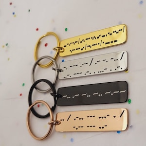 Op de afbeelding: Vier sleutelhangers met verschillende gekleurde metalen labels. Elk label heeft een andere boodschap in braille geschreven. De sleutelhangers zijn goud, zilver, zwart en roségoud.