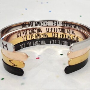 May include: Four stacked silver, rose gold, and gold bangle bracelets with the inscription "You are amazing. Keep fucking going." on each bracelet.