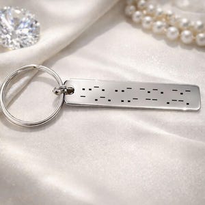 May include: A silver-colored rectangular keychain with Morse code engraving. The keychain is attached to a large silver ring. The background features a large diamond and a string of pearls, all resting on a white, silky fabric.
