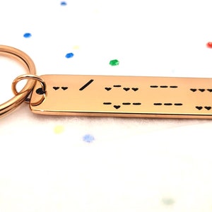 May include: A rose gold keychain with a rectangular tag engraved with a message in braille. The tag is attached to a split ring.