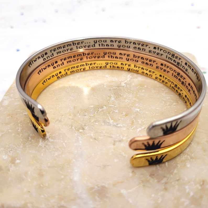 May include: Three stacked metal bracelets with a crown design and the inscription "Always remember... you are braver, stronger, smarter and more loved than you can ever imagine."