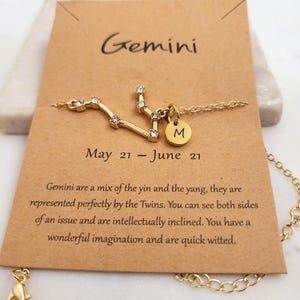 May include: Gold necklace with a Gemini constellation charm and a small letter 'M' charm. The necklace is displayed on a card with the text 'Gemini, May 21 - June 21' and a description of the Gemini zodiac sign.