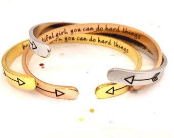 May include: Three gold, rose gold, and silver bangle bracelets with an arrow design and the inscription "beautiful girl, you can do hard things, you can do hard things".