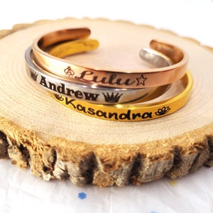 May include: Three gold, silver, and rose gold bracelets with engraved names "Lulu", "Andrew", and "Kasandra". Each name is accompanied by a crown and other decorative symbols.