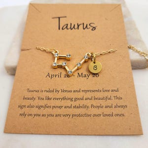 May include: Gold necklace with a Taurus constellation charm and a letter B charm. The necklace is on a brown card with the text "Taurus" and "April 20 - May 20".