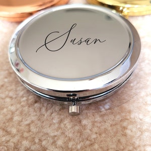 May include: A silver compact mirror with a hinged lid and a silver button on the bottom. The mirror has a personalized inscription that reads "Susan".