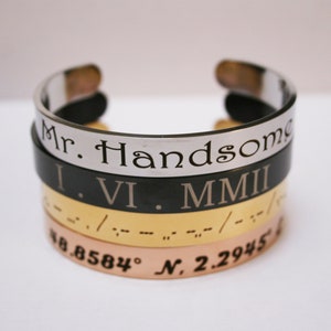 Op de afbeelding: Een stapel van vier armbanden van zilver, zwart, goud en roségoud met gegraveerde tekst. De bovenste armband heeft de tekst "Mr. Handsome", de tweede armband heeft de tekst "I. VI. MMIII", de derde armband heeft braille-tekst en de onderste armband heeft de tekst "38.8584° N, 2.2945".