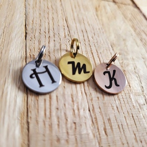 May include: Three round metal pet ID tags with engraved letters. The tag on the left is silver with the letter "H" engraved on it. The middle tag is gold with the letter "M" engraved on it. The tag on the right is rose gold with the letter "K" engraved on it.