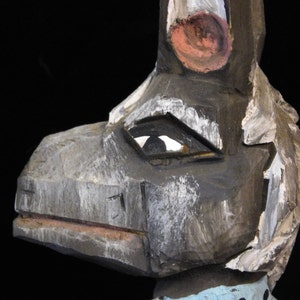 May include: A wooden carved sculpture of a stylized animal with a large beak, painted in shades of gray, white, and pink. The sculpture has a single eye with a black pupil.