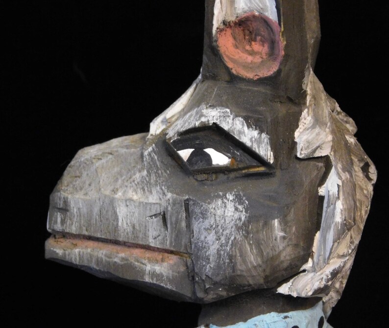 May include: A wooden carved sculpture of a stylized animal with a large beak, painted in shades of gray, white, and pink. The sculpture has a single eye with a black pupil.