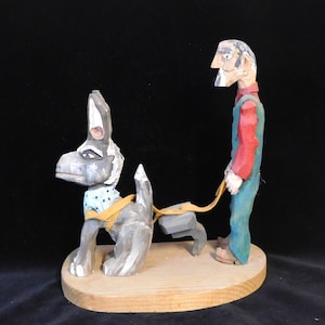 May include: A hand-carved wooden figurine of a man in overalls walking a dog. The man is painted in red, green, and white, and the dog is painted in black, white, and blue.