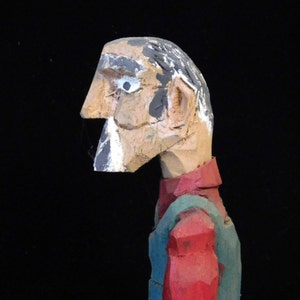 May include: A wooden carved figure of a person with a red and green shirt. The figure has a painted face with a white eye and a black beard.