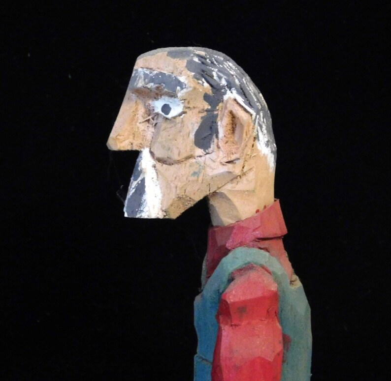 May include: A wooden carved figure of a person with a red and green shirt. The figure has a painted face with a white eye and a black beard.