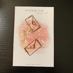 Op de afbeelding: Twee roségouden plannerclips in envelopvorm met hartdetails op een witte kaart. De kaart heeft een roze aquarelachtergrond met gouden spikkels en de tekst "PLANNER CLIP happy mail" en "QUEEN OF CENTS".