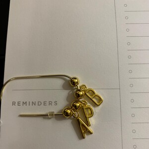 May include: A gold-toned metal safety pin brooch featuring three small gold spherical beads and three dangling gold letter charms spelling out the initials A, P, and B. The brooch includes a clear plastic stopper on the pin end.
