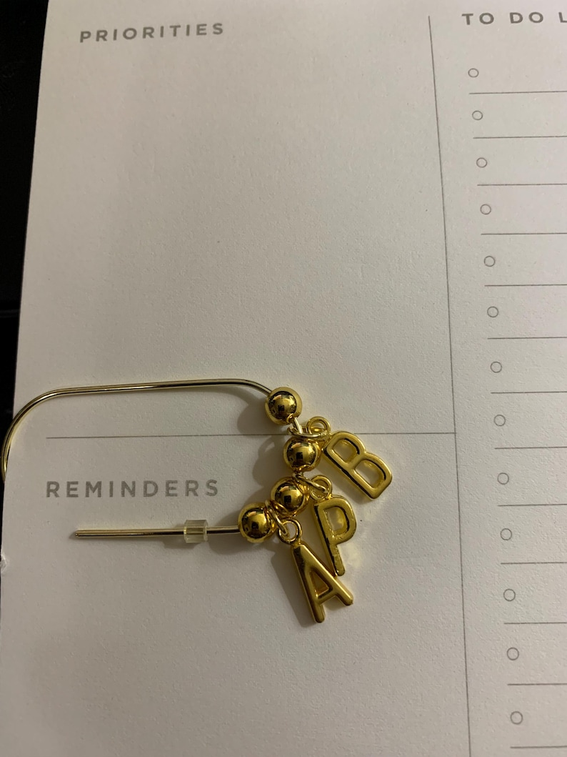 May include: A gold-toned metal safety pin brooch featuring three small gold spherical beads and three dangling gold letter charms spelling out the initials A, P, and B. The brooch includes a clear plastic stopper on the pin end.