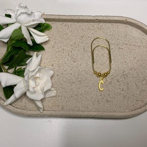 May include: A beige speckled oval tray featuring a gold-coloured paperclip-style charm with a letter "C" pendant. The charm has a small cluster of beads. Two white gardenia flowers and green leaves are arranged on the tray.