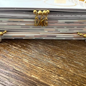 May include: Three gold-toned metal page markers attached to the top edge of a stack of paper. The center marker features three dangling letter charms spelling DKG, while the left marker has a white pearl bead and the right has a clear crystal.