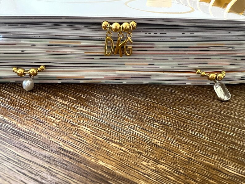 May include: Three gold-toned metal page markers attached to the top edge of a stack of paper. The center marker features three dangling letter charms spelling DKG, while the left marker has a white pearl bead and the right has a clear crystal.