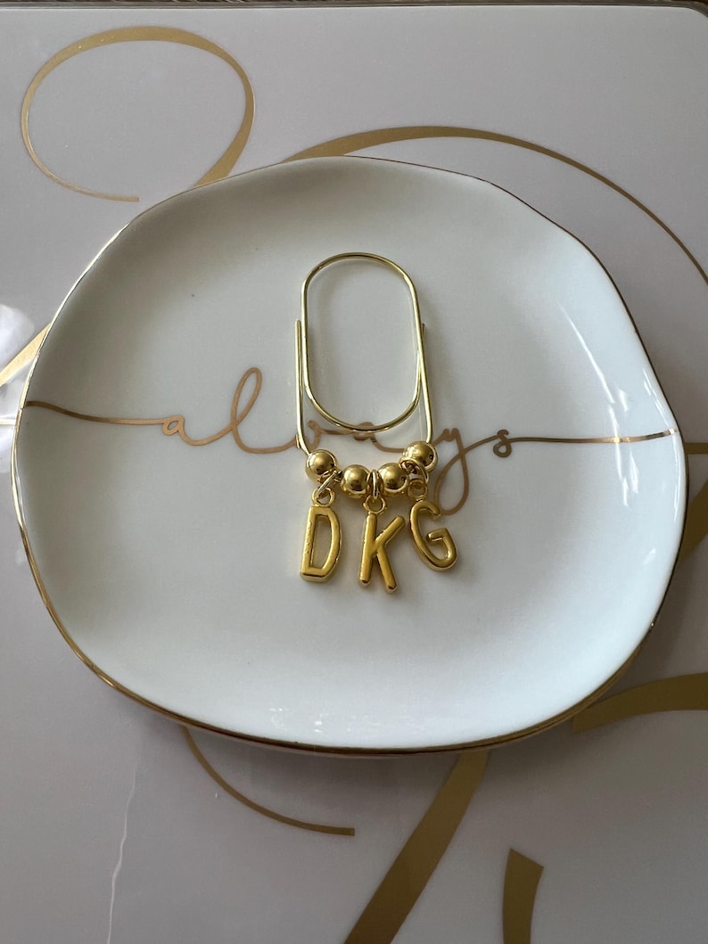 May include: A gold-toned metal paperclip-style bookmark featuring three small gold beads and three dangling gold letter charms spelling DKG. The accessory has a polished metallic finish and a simple, elongated oval shape for securing pages.