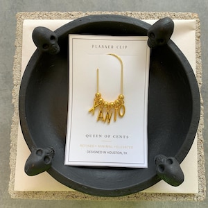 May include: A gold-toned metal planner clip featuring a U-shaped wire adorned with small gold beads and dangling letter charms spelling XANIO. The clip is attached to a white rectangular display card labelled PLANNER CLIP, QUEEN OF CENTS, REFINED MINIMAL ELEVATED, and DESIGNED IN HOUSTON, TX.