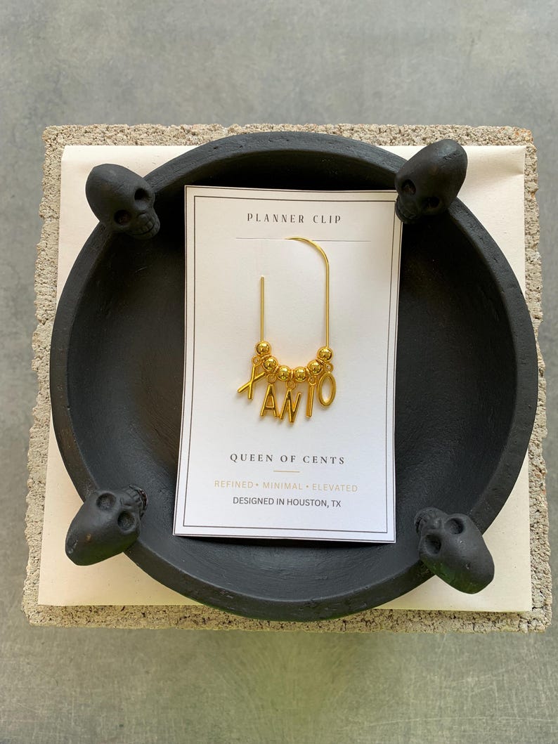 May include: A gold-toned metal planner clip featuring a U-shaped wire adorned with small gold beads and dangling letter charms spelling XANIO. The clip is attached to a white rectangular display card labeled PLANNER CLIP, QUEEN OF CENTS, REFINED MINIMAL ELEVATED, and DESIGNED IN HOUSTON, TX.