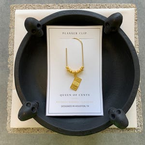 May include: A gold-toned metal planner clip featuring a small dangling rectangular charm shaped like a 100 dollar bill, accented with a row of tiny gold beads. The clip is attached to a white display card with the text PLANNER CLIP, QUEEN OF CENTS, REFINED MINIMAL ELEVATED, and DESIGNED IN HOUSTON, TX.