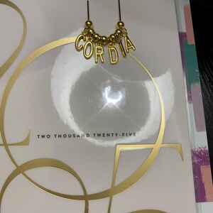 May include: A gold-toned metal paperclip bookmark featuring a curved top and a dangling charm section. The charm consists of a row of small gold beads interspersed with individual gold letter charms that spell out the name CORDIA in capital letters.
