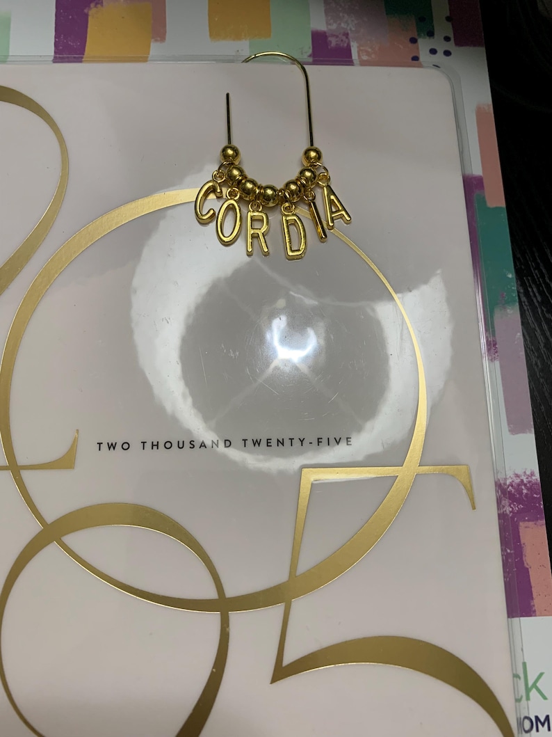 May include: A gold-toned metal paperclip bookmark featuring a curved top and a dangling charm section. The charm consists of a row of small gold beads interspersed with individual gold letter charms that spell out the name CORDIA in capital letters.