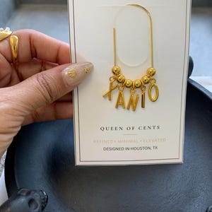 May include: A gold-toned metal planner clip attached to a white display card. The clip features a curved wire design adorned with five small gold beads and four dangling gold letter charms spelling the name XANI. The card reads PLANNER CLIP, QUEEN OF CENTS, REFINED, MINIMAL, ELEVATED, and DESIGNED IN HOUSTON, TX.