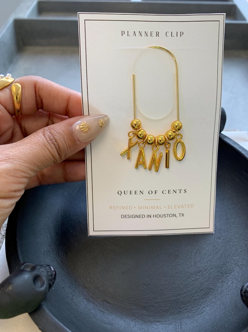 May include: A gold-toned metal planner clip attached to a white display card. The clip features a curved wire design adorned with five small gold beads and four dangling gold letter charms spelling the name XANI. The card reads PLANNER CLIP, QUEEN OF CENTS, REFINED, MINIMAL, ELEVATED, and DESIGNED IN HOUSTON, TX.