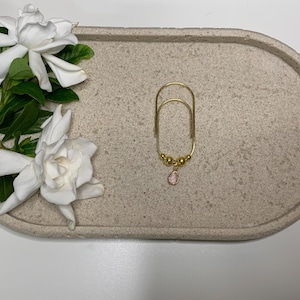 May include: A gold-colored earring holder with a pink gemstone pendant and small gold beads, resting on a light gray oval tray. White gardenia flowers and green leaves are arranged on the tray's edge.