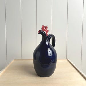 May include: A navy blue ceramic pitcher with a red rooster head design on top. The pitcher has a handle and a glossy finish. The rooster's comb and beak are red, contrasting with the dark blue body.